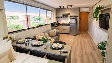 A modern hotel room with a dining area, kitchenette, and a large window providing a view of the outdoors.