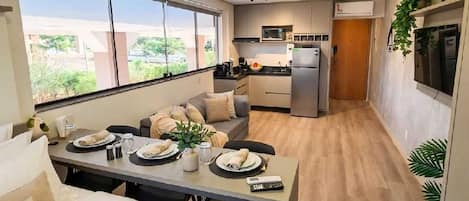 A modern hotel room with a dining area, kitchenette, and a large window providing a view of the outdoors.