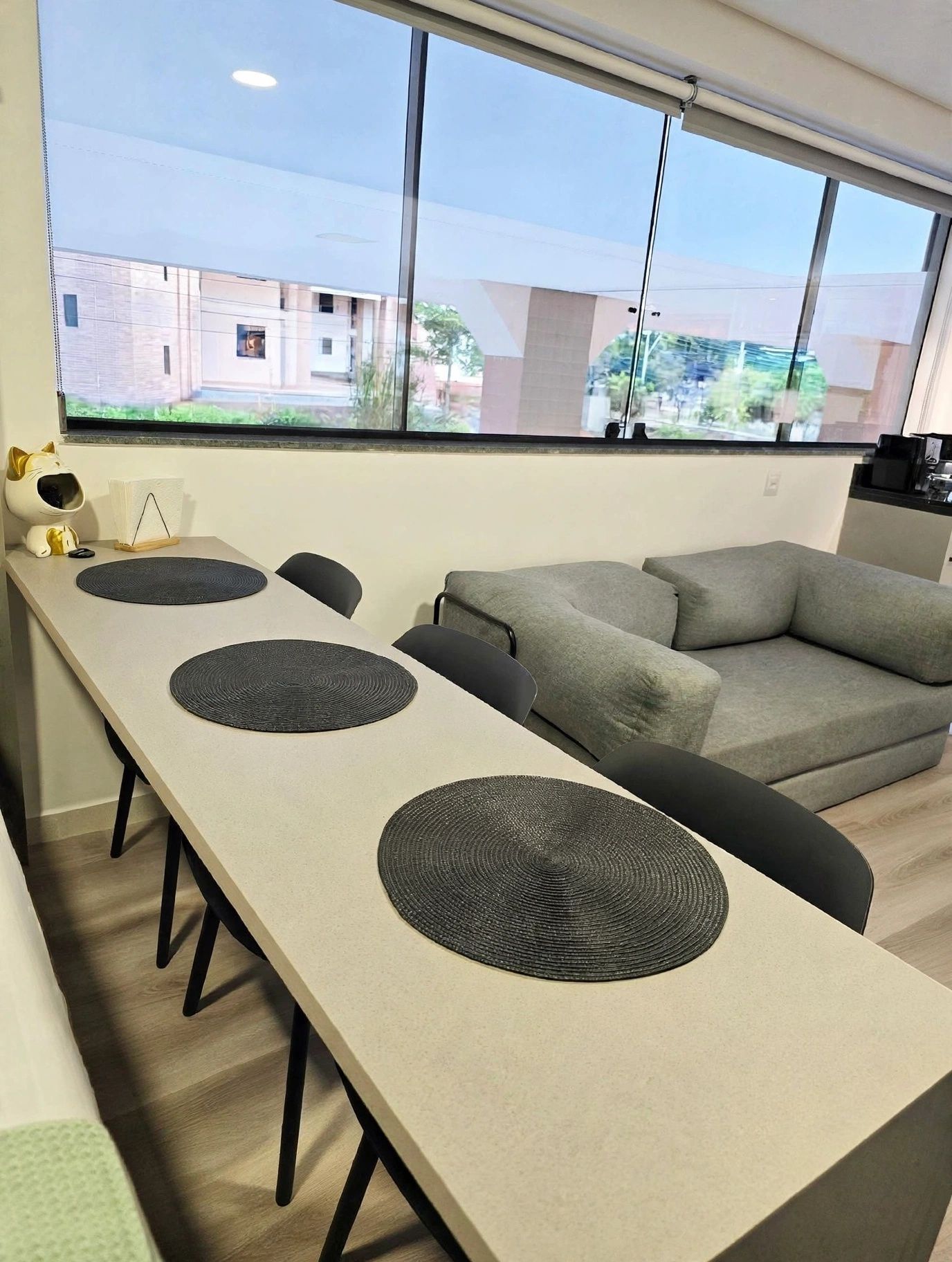 Uma sala de estar moderna com uma mesa comprida, dois sofás e uma janela grande com vista para prédios.