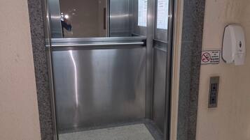 Um elevador com portas de aço inoxidável, um dispensador de álcool em gel e avisos de segurança na parede.