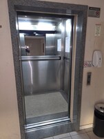 Um elevador com portas de aço inoxidável, um dispensador de álcool em gel e avisos de segurança na parede.