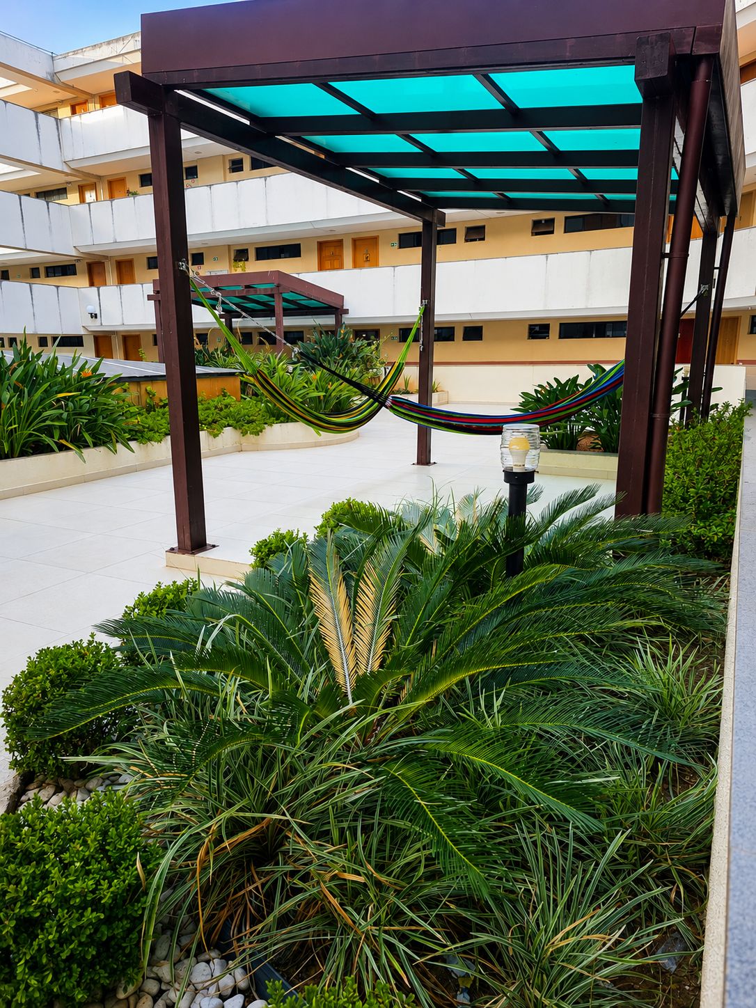 A covered outdoor area with a hammock, palm plants, and a building with multiple floors in the background.