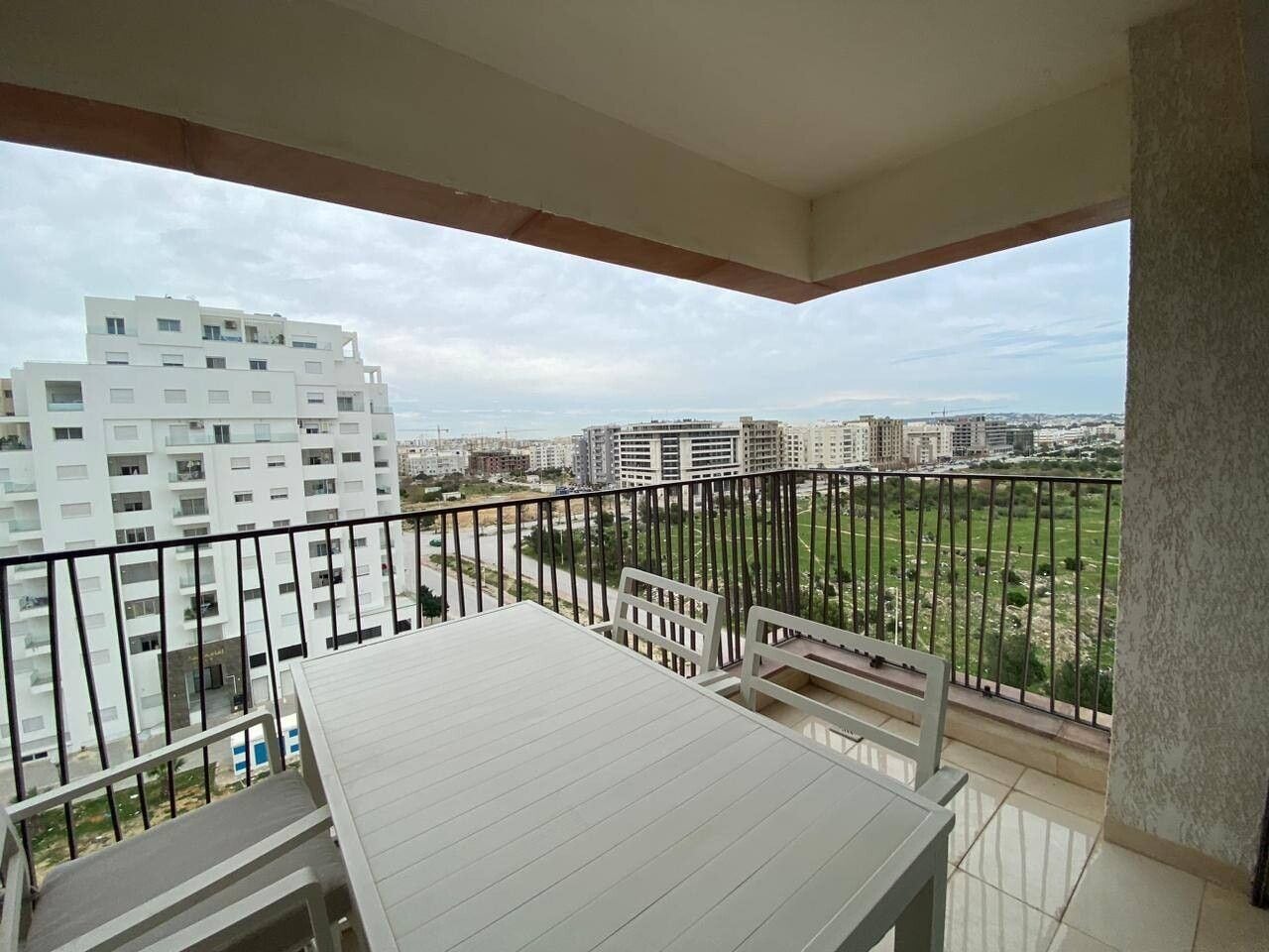 Un balcon avec une table et des chaises, donnant sur un paysage urbain avec des immeubles et un parc.