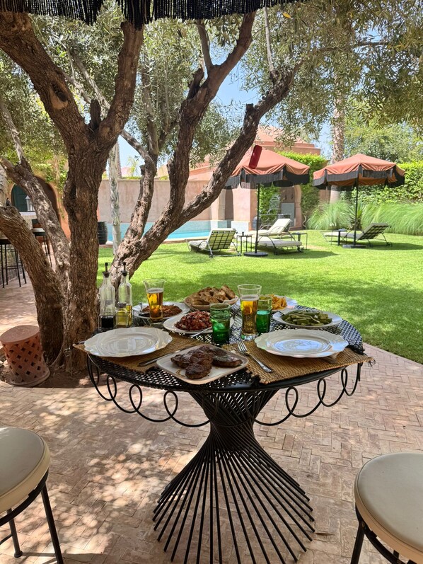 An outdoor dining area with a table set for a meal, surrounded by trees and a view of a pool and lounge chairs. - Villa in Marrakech, with a private pool (Marrakech)