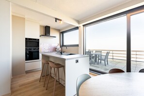 A modern kitchen with an island, bar stools, and a dining area with a table and chairs. - Penthouse by the Sea With Stunning Rooftop Views (Blankenberge)