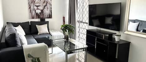 Un salon moderne avec un canapé noir, une table basse en verre, un grand téléviseur et un tableau représentant un lion.