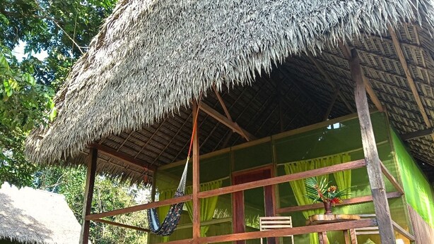 Una cabaña con techo de paja, estructura de madera, una hamaca y una ventana con cortinas.