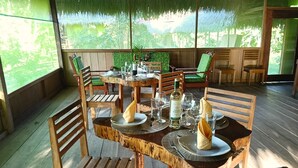 Una mesa de comedor de madera dispuesta con platos, vasos y una botella, rodeada de sillas en un espacio exterior cubierto con vistas a la vegetación.