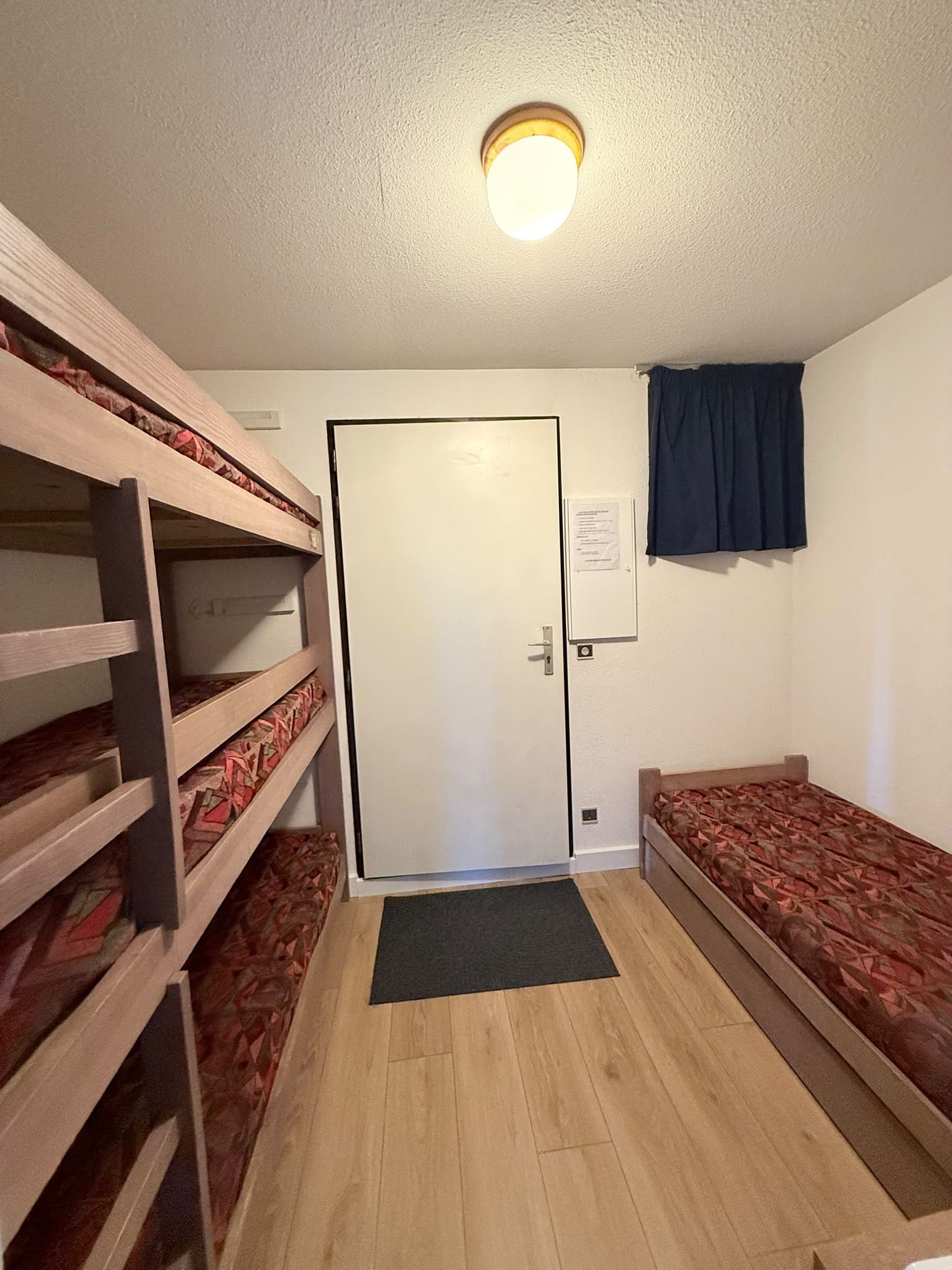 A room with bunk beds, a door, a small window with curtains, and a light fixture on the ceiling.