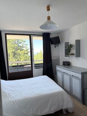 A hotel room with a bed, a desk with a computer, a TV, and a view of trees through the window. - Studio for 4 – Résidence Les Marmottes – 1750 Chamrousse (Chamrousse)