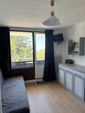 A room with a bed, a TV mounted on the wall, a small kitchenette, and a sliding glass door leading to an outdoor area. - Studio for 4 – Résidence Les Marmottes – 1750 Chamrousse (Chamrousse)