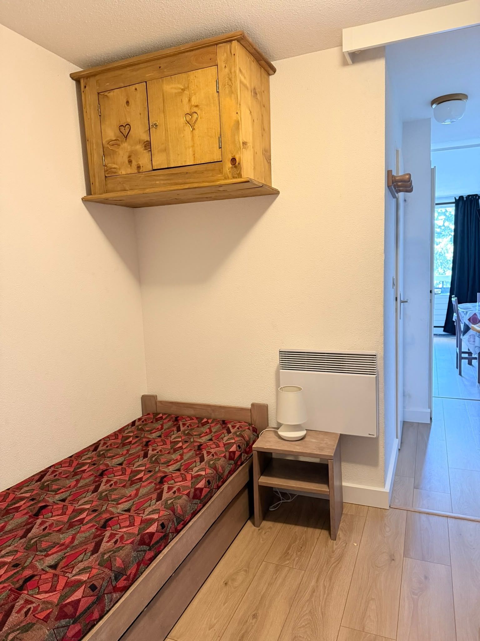 Un dormitorio con una cama, una mesita de noche pequeña, un calentador y un armario de madera sobre la cama.