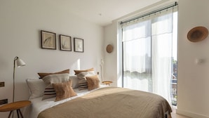 Une chambre à coucher avec un grand lit, une table de chevet, une lampe sur pied et une fenêtre ornée de rideaux légers.