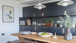 Une cuisine moderne avec des meubles sombres, des suspensions blanches et une table à manger en bois sur laquelle on a disposé un saladier de fruits et quelques livres.