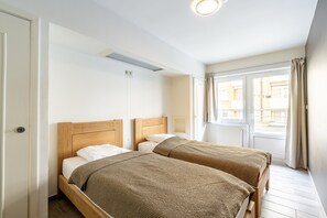 Une chambre à coucher avec deux lits en bois, des murs blancs, une fenêtre avec des rideaux et un radiateur.
