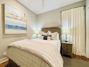 A bedroom with a bed, nightstands, lamps, a painting, and a window with curtains.