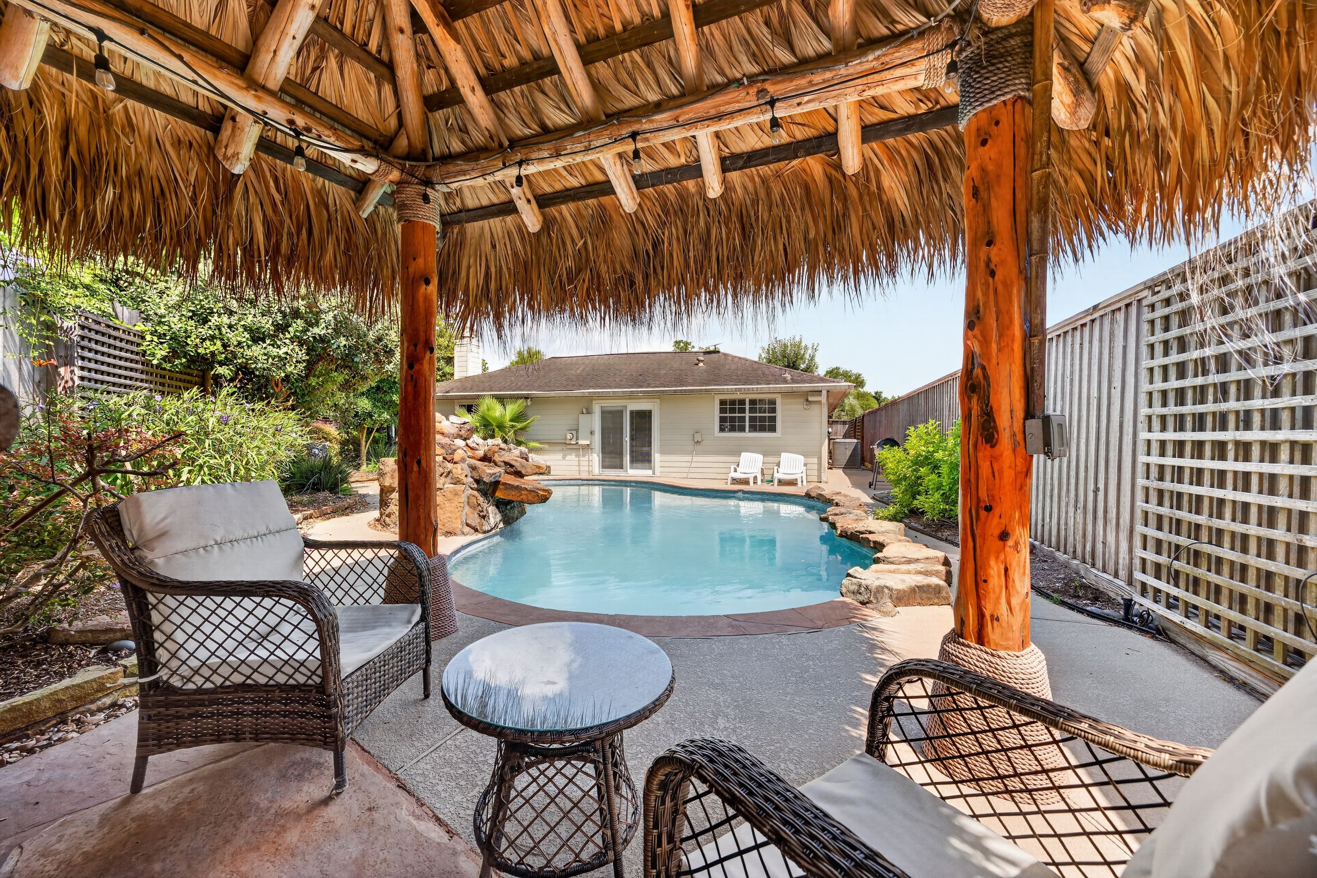 A pool area with a thatched roof, wicker furniture, and a view of a house in the background.