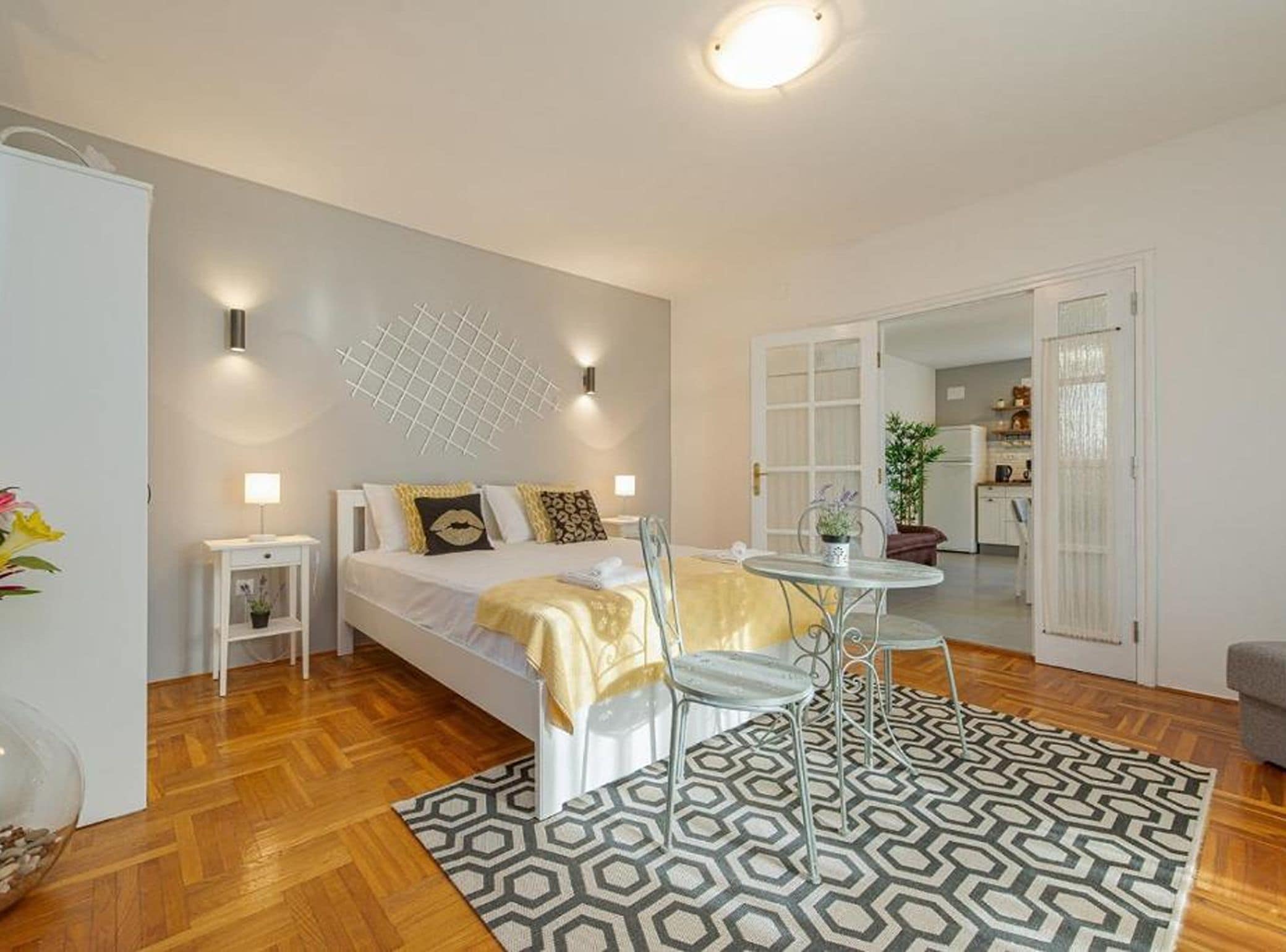 A modern bedroom with a bed, bedside tables, a small table, and a chair. There is a patterned rug on the floor.