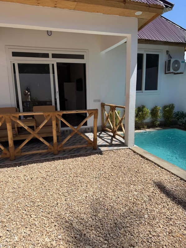 Uma área à beira da piscina com mesa e cadeiras de madeira, um espaço de estar coberto e um edifício com fachada branca e janela com unidade de ar condicionado.