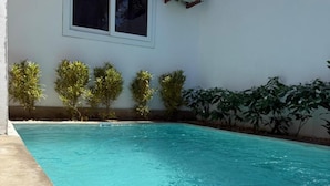 Una piscina con agua azul cristalina, rodeada por un edificio blanco con una ventana y un equipo de aire acondicionado.