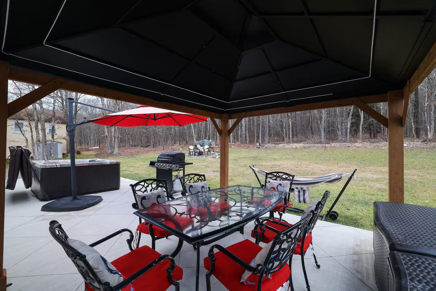 Un patio exterior con una mesa de vidrio, sillas rojas acolchadas, una parrilla y un paraguas rojo.