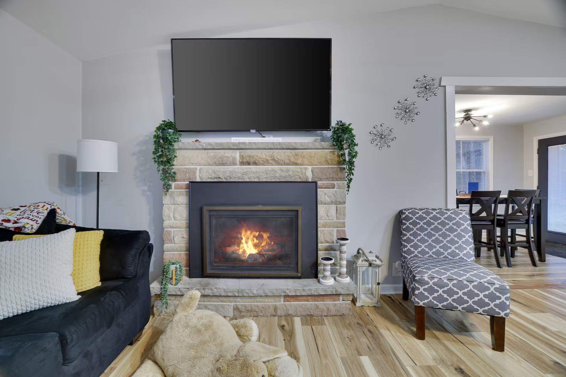 Een woonkamer met een open haard, een flatscreen televisie erboven, een bank, een gestreept fauteuil, en een eethoek op de achtergrond.