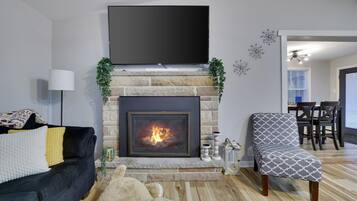 Een woonkamer met een open haard, een flatscreen televisie erboven, een bank, een gestreept fauteuil, en een eethoek op de achtergrond.