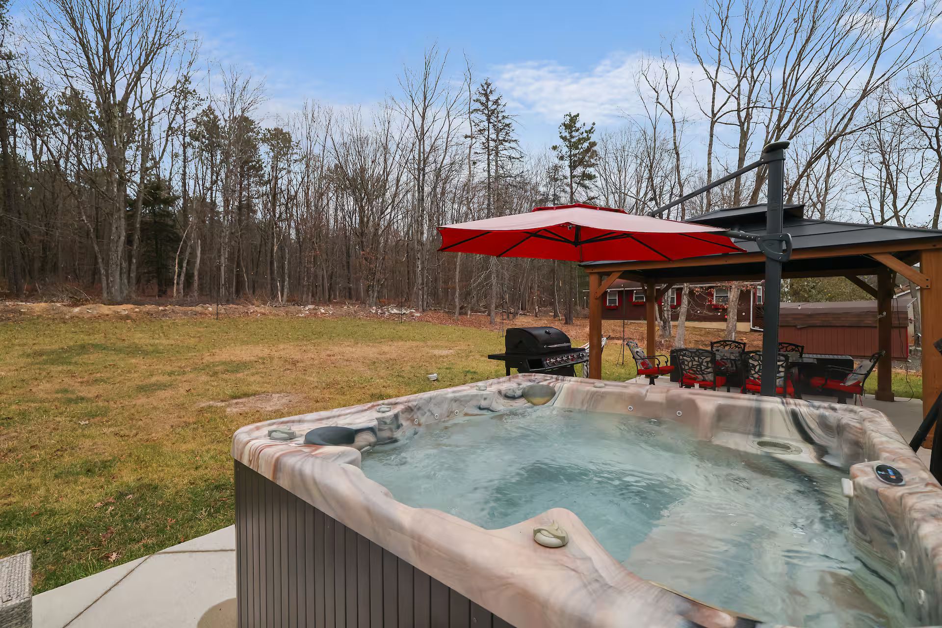 Un jacuzzi con cubierta de madera y un paraguas rojo, rodeado de césped y árboles.