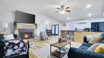 A modern living room with a fireplace, a ceiling fan, and a kitchen area in the background.