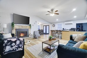 A modern living room with a fireplace, a ceiling fan, and a kitchen area in the background.