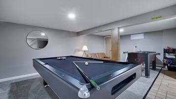 A pool table with balls, a sofa, a lamp, and a foosball table in a room with grey walls and a mirror.
