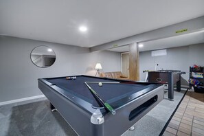 A pool table with balls, a sofa, a lamp, and a foosball table in a room with grey walls and a mirror.