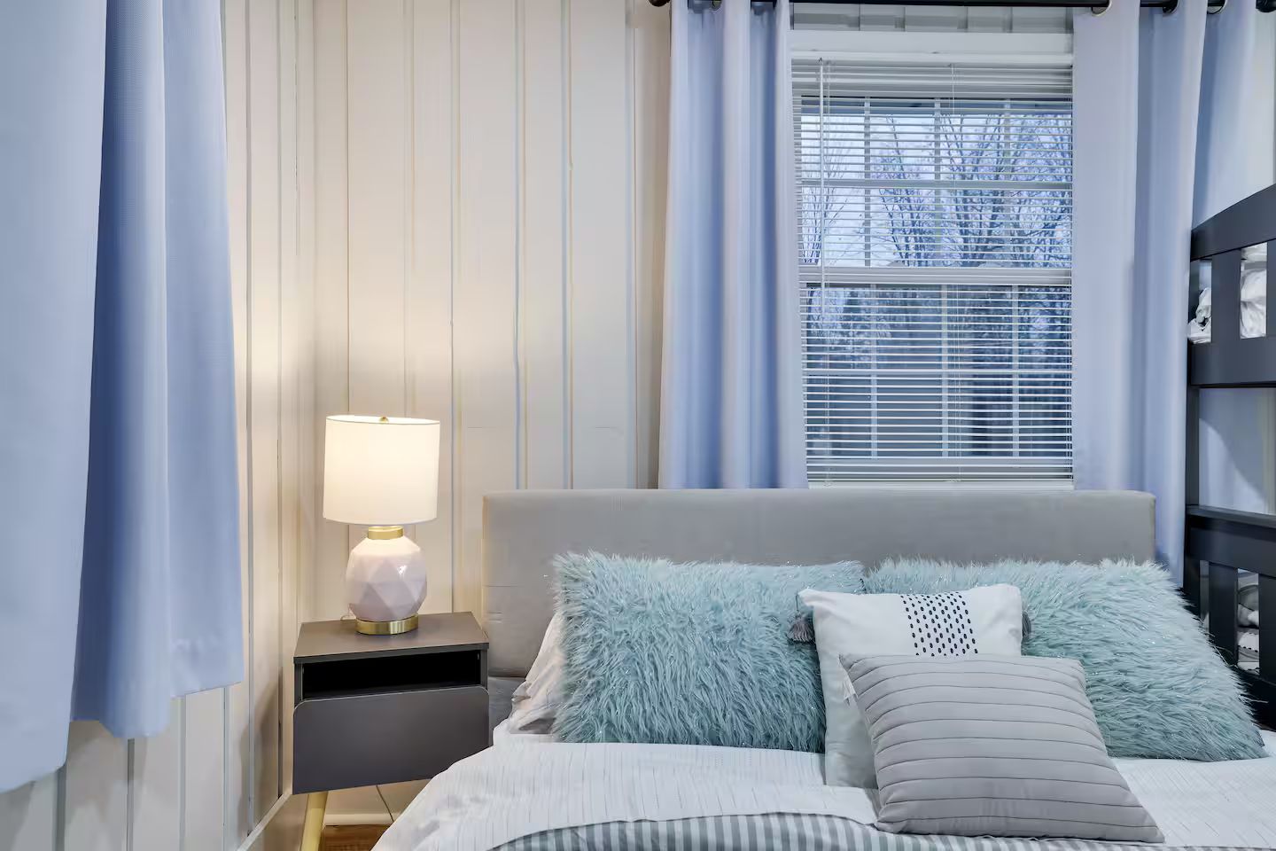A bedroom with a bed, a nightstand with a lamp, and a window with blinds.