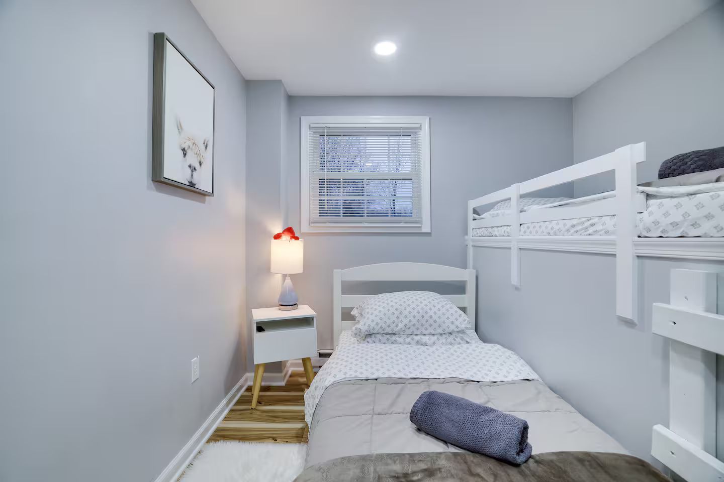 Una habitación pequeña con una cama individual, una mesita de noche, una lámpara, una ventana con persianas y una cama nido a la derecha.