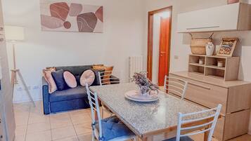 A dining area with a table and chairs, a sofa, a lamp, and a wall-mounted shelf with decorative items.