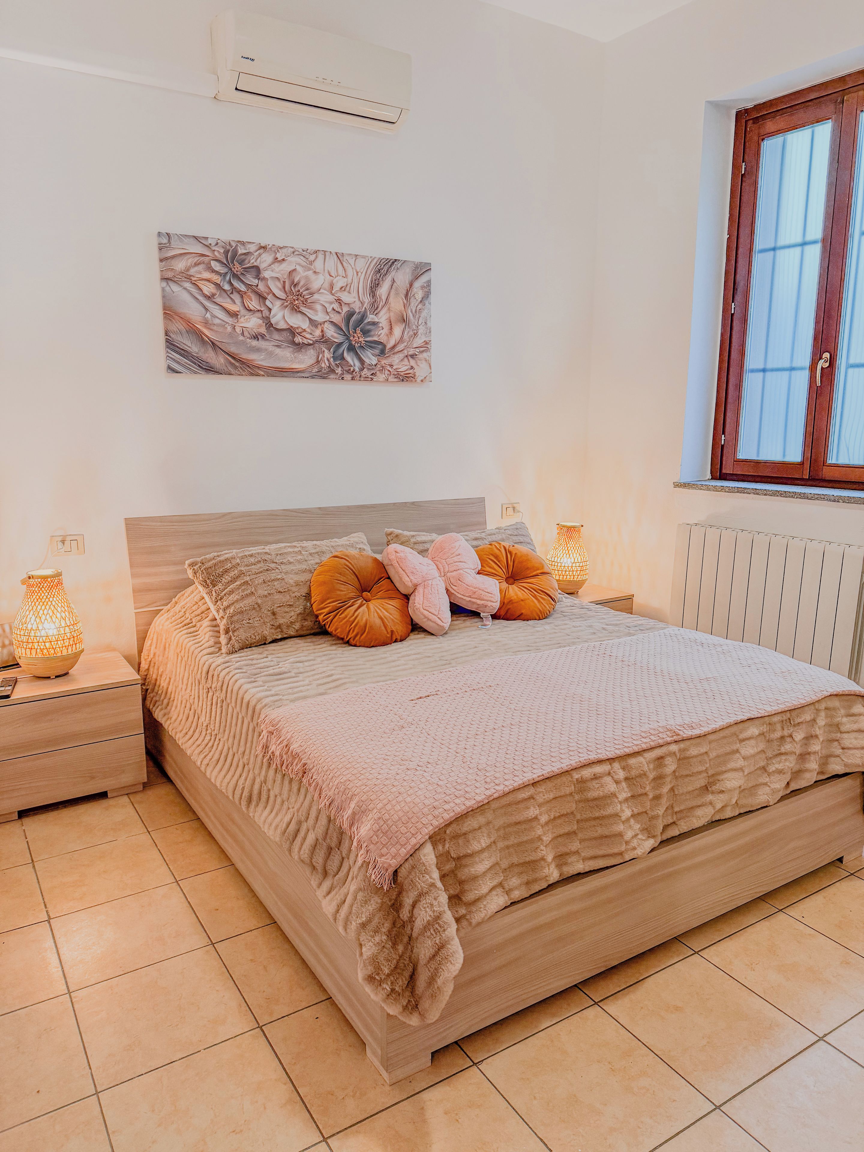 A bedroom with a wooden bed, a nightstand with a lamp, a wall-mounted air conditioner, and a window with a wooden frame.