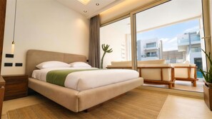 Ein modernes Schlafzimmer mit einem großen Bett, einer Couch, einer Kakteenpflanze und Blick auf ein Gebäude durch eine Glastür.