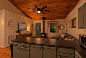 A cozy interior with a wooden ceiling, a central fireplace, a dining area, and a living space with a sofa and cushions.