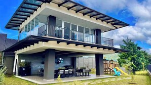 Een modern huis met twee verdiepingen, een balkon, een patio met tuinmeubilair en een speelruimte voor kinderen.