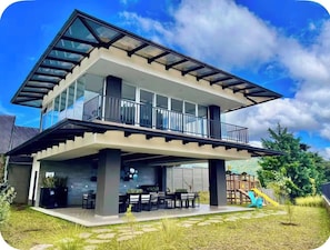 Une maison moderne à deux étages dotée d’un balcon, d’une terrasse avec mobilier d’extérieur et d’un espace de jeux pour enfants.