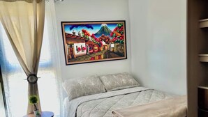 Een slaapkamer met een bed, een schilderij, een klein tafeltje met een plant en een raam met gordijnen.