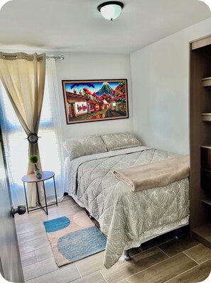 Une chambre à coucher avec un lit, un tableau, une petite table avec une plante et une fenêtre avec des rideaux.