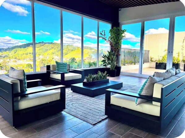 A modern living room with a sofa, armchairs, and a coffee table, featuring a large window with a view of a green landscape.