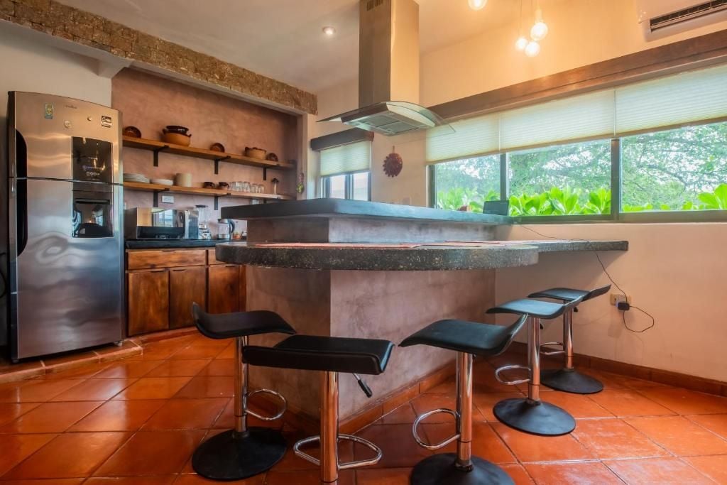 Cocina con isla integrada, taburetes de bar, refrigerador y ventana con vista a follaje.