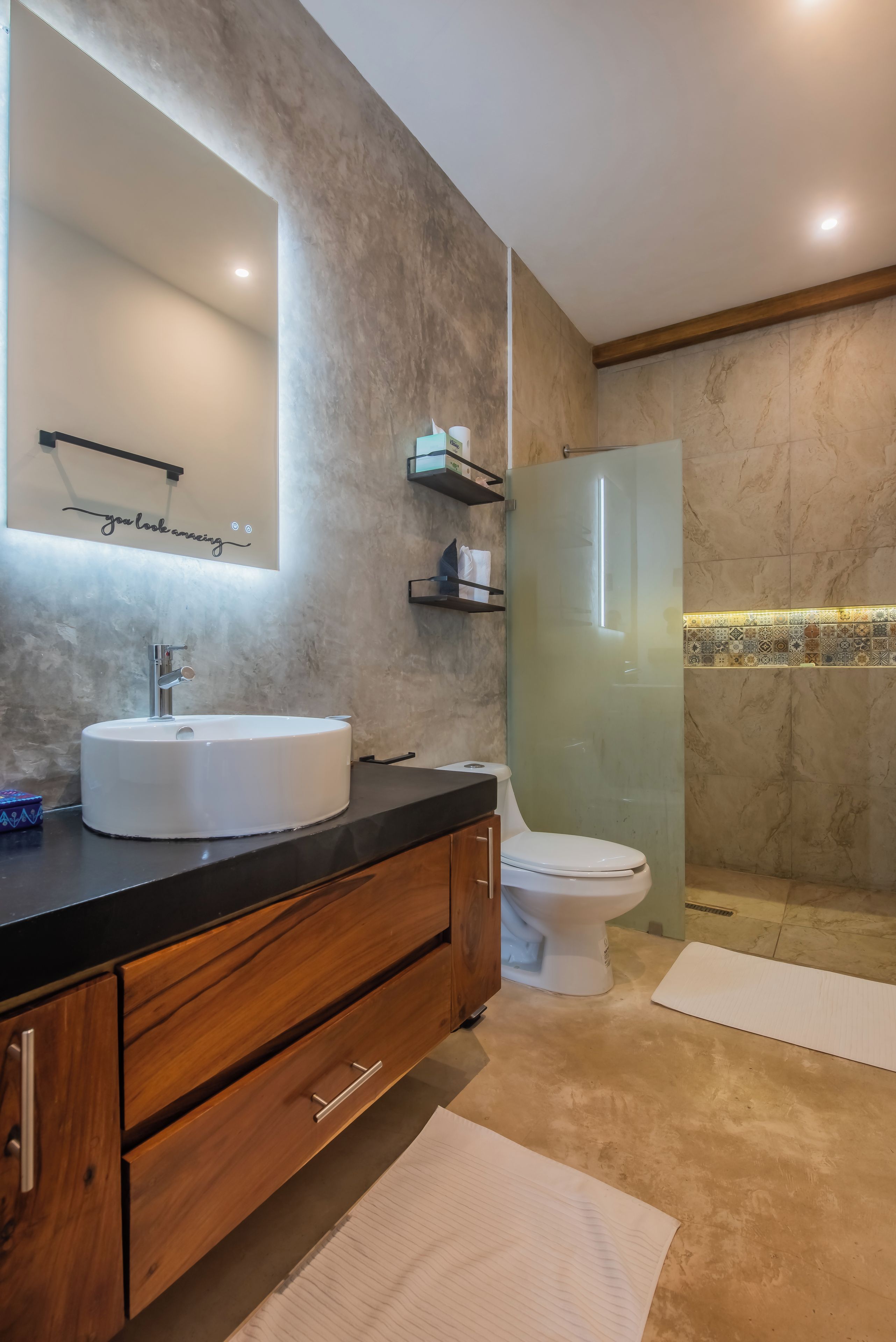 Un baño moderno con pared de mármol, un espejo grande, un lavamanos blanco, un inodoro y una zona de ducha.