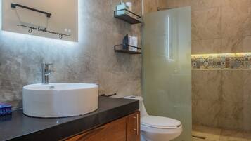 Un baño moderno con pared de mármol, un espejo grande, un lavamanos blanco, un inodoro y una zona de ducha.