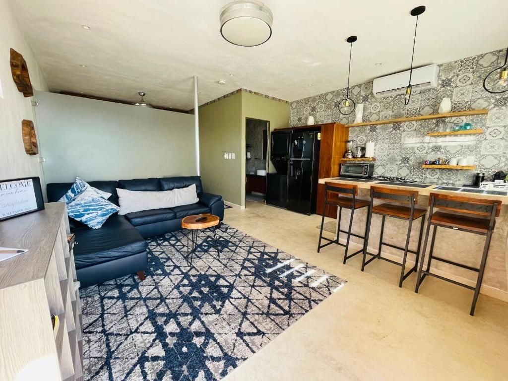 Una sala de estar moderna con un sofá negro, una mesa de centro y un tapete estampado. Junto a la sala se encuentra una cocina con microondas, refrigerador y taburetes de bar.