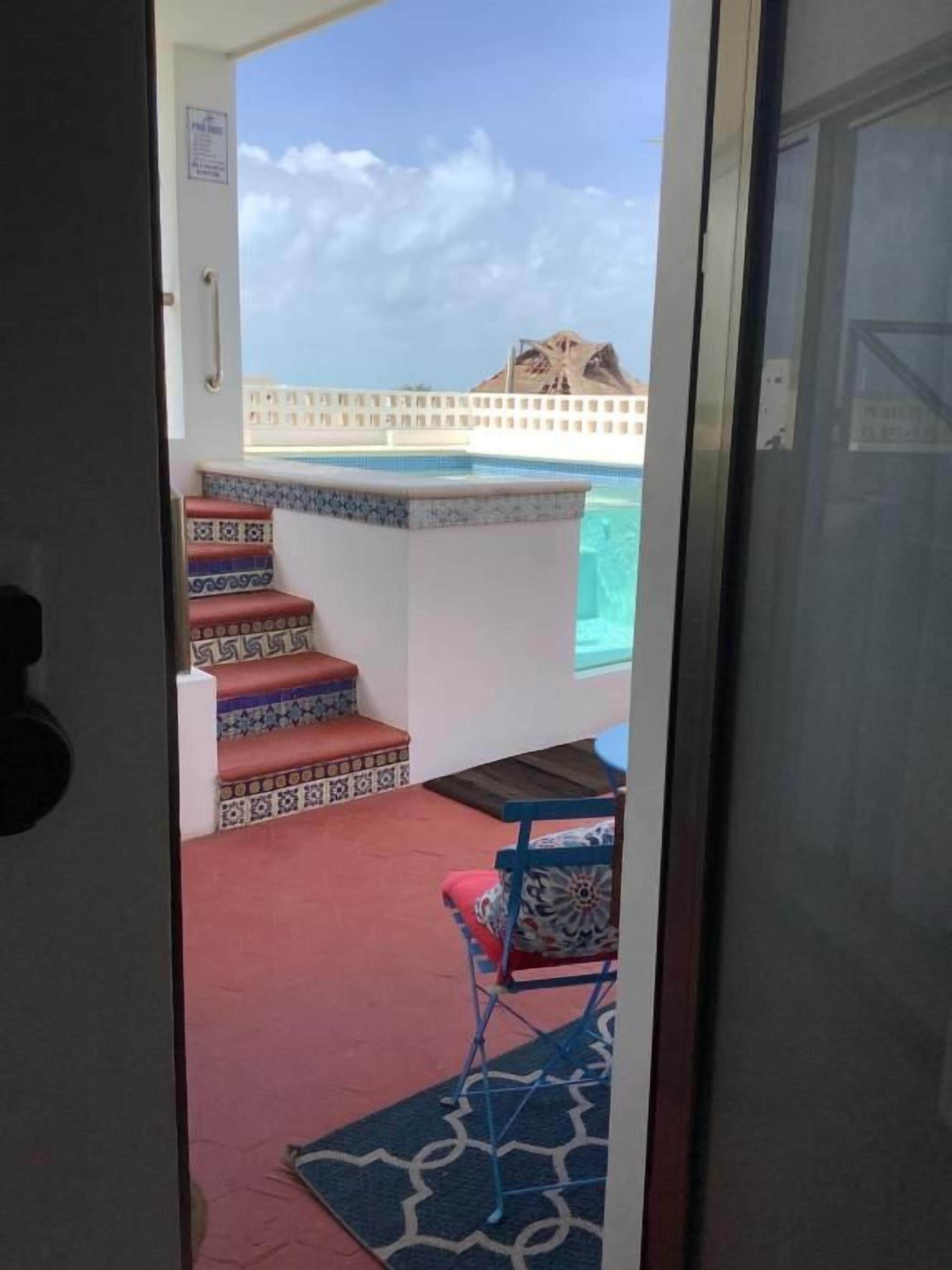Vista a través de una puerta de un área de piscina con una escalera que baja a la piscina, una silla y un tapete estampado.