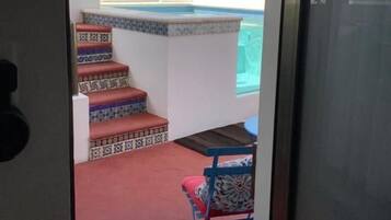 Vista a través de una puerta de un área de piscina con una escalera que baja a la piscina, una silla y un tapete estampado.