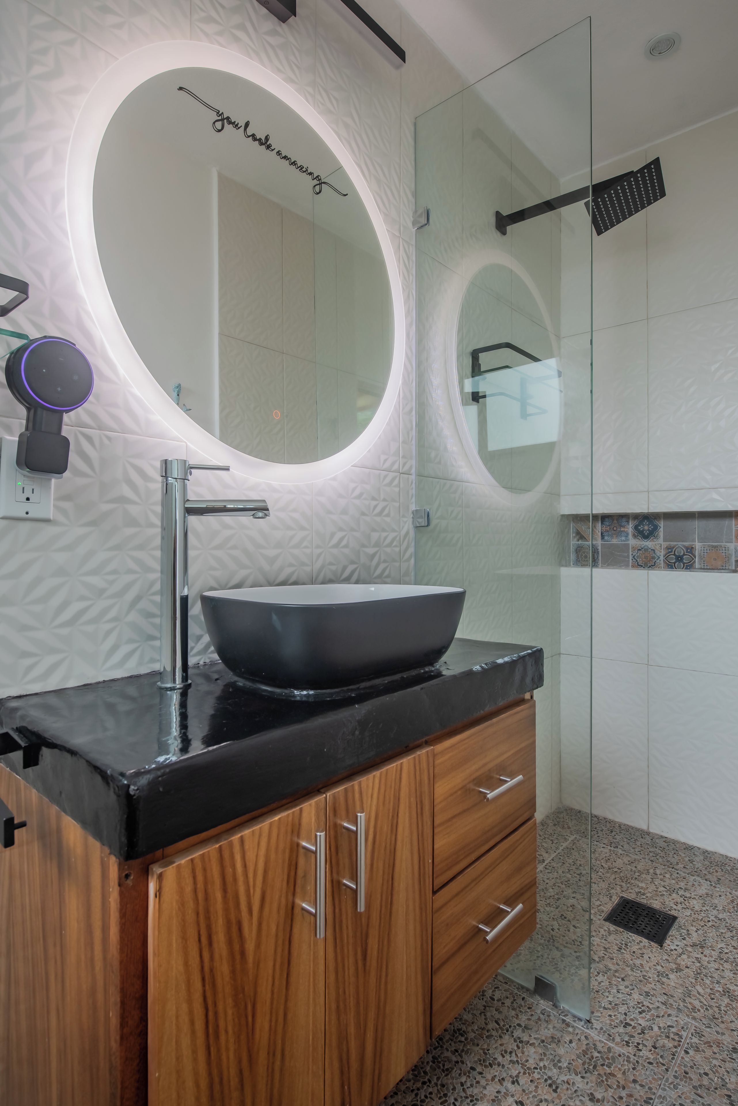 Un baño moderno con un espejo redondo, un lavabo de diseño negro, armarios de madera y un box de ducha de vidrio.